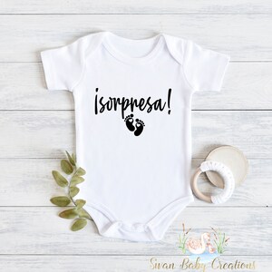 May include: White baby onesie with black text that says "¡sorpresa!" and a pair of black baby footprints.
