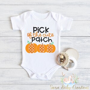 May include: White baby onesie with the text "Pick of the cute Patch" in black and orange glitter. Three orange pumpkins with white polka dots are below the text. A wooden baby teether is next to the onesie.