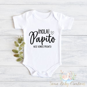 May include: White baby onesie with black text that reads "¡HOLA! Papito NOS VEMOS PRONTO".