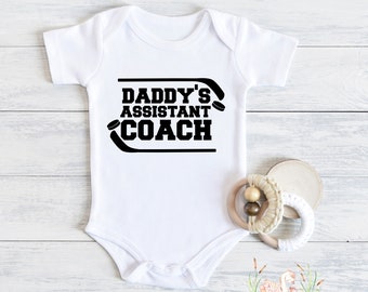 May include: White baby onesie with the text "Daddy's Assistant Coach" in black. The design includes hockey sticks and pucks. A wooden baby teether with a cream-colored knitted ring and wooden beads is next to the onesie.