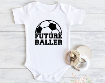 May include: White baby onesie with a black soccer ball graphic and the text "FUTURE BALLER". A wooden teething ring and a wooden disc are next to the onesie. The onesie is on a white wooden surface.
