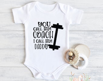 May include: White baby onesie with black text that reads "You call him coach I call him daddy" with a black dumbbell graphic.