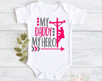 May include: White baby onesie with pink text that reads "My Daddy My Hero" with a silhouette of a lineman climbing a utility pole.