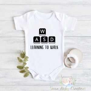 May include: White baby onesie with black text and a graphic of computer keyboard keys that say "W, A, S, D" and "Learning to Walk".