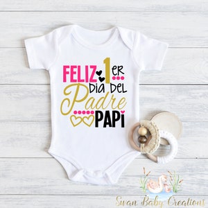 May include: White baby onesie with the text "Feliz 1er dia del Padre Papi" in pink, gold, and black lettering with pink hearts.