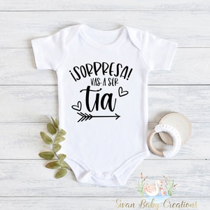 May include: White baby onesie with black text that reads "¡Sorpresa! Vas a ser tía" with a black arrow pointing to the right and two black hearts.