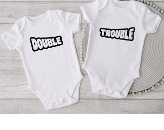 May include: Two white baby onesies with black text. One onesie says "DOUBLE" and the other says "TROUBLE". The text is in a bold, rounded font with a black outline. The onesies are on a light wood-grain surface.
