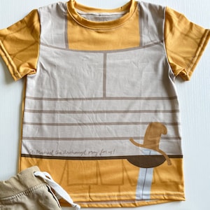 Peut inclure: Un t-shirt à manches courtes jaune et beige avec un graphique de chevalier et le texte "St. Michael the Archangel pray for us!" Le t-shirt est posé sur une surface blanche avec un pantalon marron et des chaussures marron.