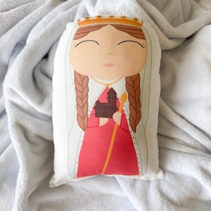 Puede incluir: Un juguete de peluche de una mujer con un vestido rojo y una corona, sosteniendo un pequeño edificio en su mano. El juguete tiene una cara sonriente y cabello castaño en trenzas.