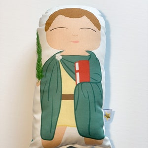 May include: A plush toy of a saint holding a book and a palm branch. The saint is wearing a green robe and a yellow tunic. The toy is made by "Small Folk Love".