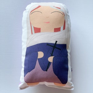 May include: A white pillow with a colorful illustration of a woman holding a cross. The woman is wearing a purple robe and a white head covering. Her hair is red and she has a gentle expression on her face.