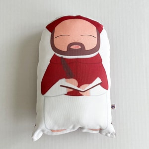 May include: A red and white plush pillow shaped like a person holding a quill and a book. The person has a brown beard and a red hat.