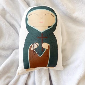 Può includere: Bambola di peluche raffigurante una figura religiosa, con un volto sereno, una veste con cappuccio verde scuro e una croce marrone. La bambola tiene un rosario e un indumento marrone. Lo sfondo è un tessuto bianco strutturato.