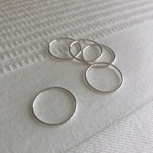 May include: Five silver rings of varying sizes are arranged on a white textured surface. The rings are simple and delicate, with a smooth, polished finish.