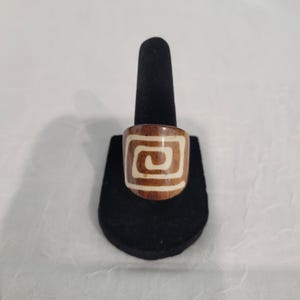 May include: A wooden ring with a brown exterior and a white spiral design. The ring is displayed on a black ring holder against a white background. The ring's design is a repeating geometric pattern.