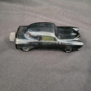 May include: A black vintage car-shaped bottle. The car has a chrome-colored cap and wheels. The car's design includes a curved roof and tail fins, suggesting a classic automobile style. The bottle is made of glass.