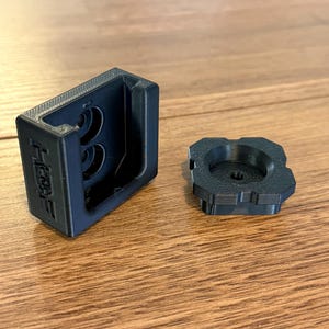 May include: Black 3D-printed plastic components, including a rectangular housing with two circular openings and a separate, circular piece. The housing has the text "HERE" on its side. The items are displayed on a wooden surface.