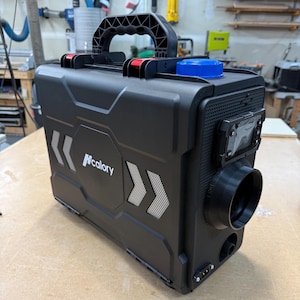 May include: A black portable heater with a handle, a blue cap, and the "Ncalory" logo. The heater has a vent and a control panel. The design includes silver chevron accents on the sides. The unit is on a wooden surface.