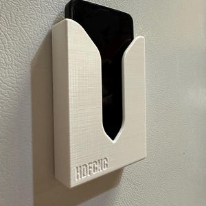 Puede incluir: Soporte de tel&eacute;fono de pl&aacute;stico blanco montado en una pared blanca. El soporte tiene un recorte para un tel&eacute;fono y el texto "HDFCNG" est&aacute; impreso en la parte inferior.