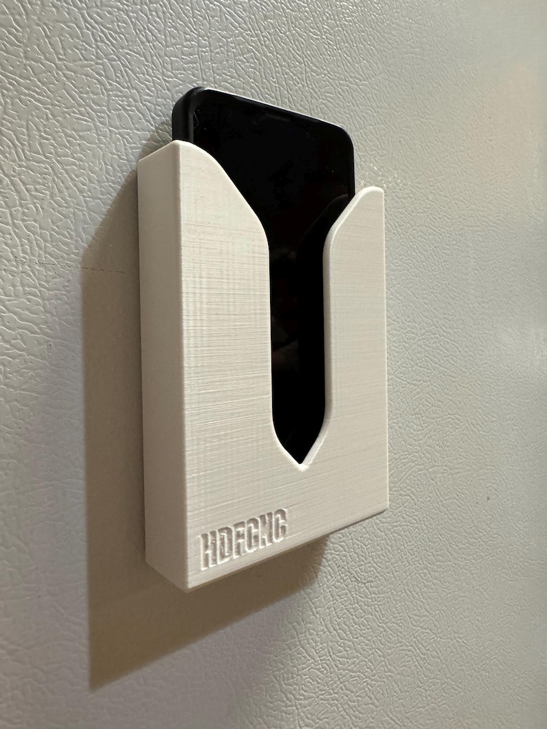 Puede incluir: Soporte de tel&eacute;fono de pl&aacute;stico blanco montado en una pared blanca. El soporte tiene un recorte para un tel&eacute;fono y el texto "HDFCNG" est&aacute; impreso en la parte inferior.