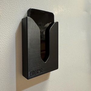 Puede incluir: Un soporte de tel&eacute;fono negro con un recorte para un tel&eacute;fono. El soporte est&aacute; montado en una pared blanca. El soporte tiene la palabra "HOUSECAR" impresa en la parte inferior.