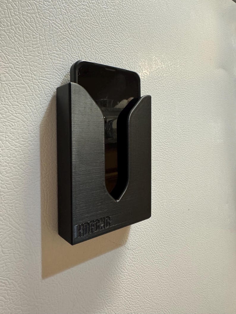 Puede incluir: Un soporte de tel&eacute;fono negro con un recorte para un tel&eacute;fono. El soporte est&aacute; montado en una pared blanca. El soporte tiene la palabra "HOUSECAR" impresa en la parte inferior.