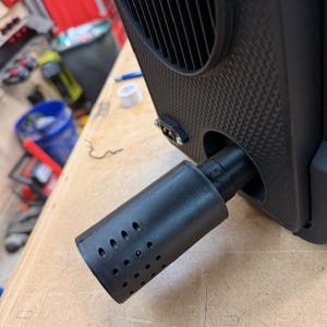 Puede incluir: Un dispositivo electrónico negro con un accesorio cilíndrico perforado. El dispositivo tiene una superficie texturizada y una rejilla de ventilación. El accesorio cilíndrico tiene múltiples agujeros pequeños. El dispositivo está sobre una superficie de madera.