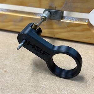 May include: A black, 3D-printed tool with a ring handle and a protruding pin. The tool is connected to a metal rod and a wooden structure. The text "HRF" is printed on the tool.