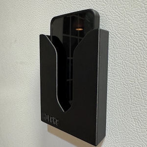 Puede incluir: Un soporte de tel&eacute;fono negro con la palabra "Hipere" impresa en la parte delantera. El soporte est&aacute; montado en una pared blanca.
