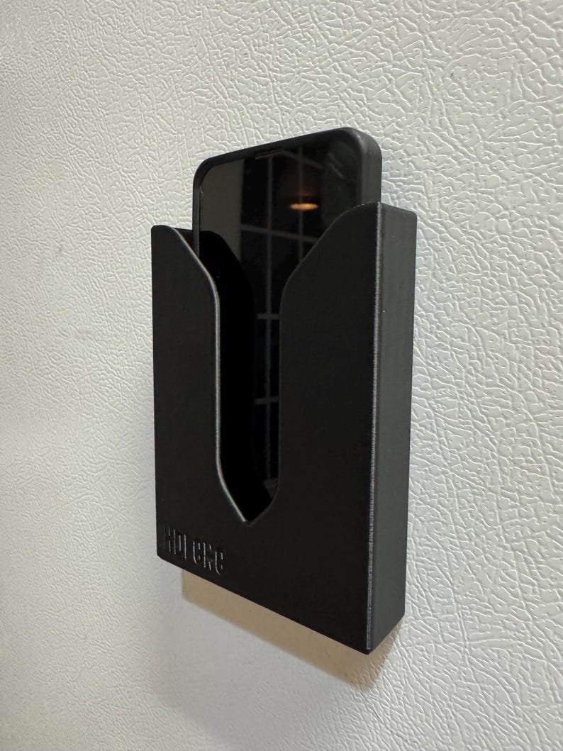 Puede incluir: Un soporte de tel&eacute;fono negro con la palabra "Hipere" impresa en la parte delantera. El soporte est&aacute; montado en una pared blanca.
