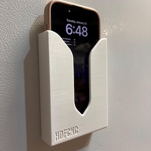 Puede incluir: Soporte para tel&eacute;fono blanco impreso en 3D con el texto "HDFCNG" en la parte inferior. Un tel&eacute;fono inteligente est&aacute; en el soporte, mostrando la hora "6:48" y la fecha "Tuesday, January 21".