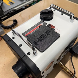 May include: A white and black industrial machine with a black handle, a black cap, and a black textured plate with the letters "HNC" on top. The machine has a gauge with red and white markings and a black nozzle.