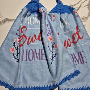 May include: Two light blue kitchen towels with the words "Sweet Home" in red and purple. The towels have a blue crochet trim and a blue crochet loop at the top. Floral designs are printed on the towels.