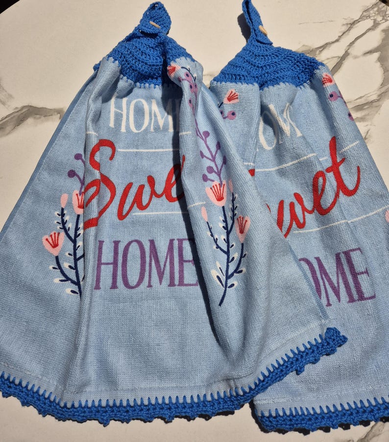 May include: Two light blue kitchen towels with the words "Sweet Home" in red and purple. The towels have a blue crochet trim and a blue crochet loop at the top. Floral designs are printed on the towels.