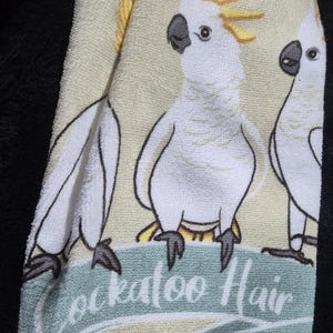 May include: A white and yellow hand towel featuring three cartoon cockatoos with the text "Cockatoo Hair Don't". The towel has a yellow crocheted trim and a loop for hanging.