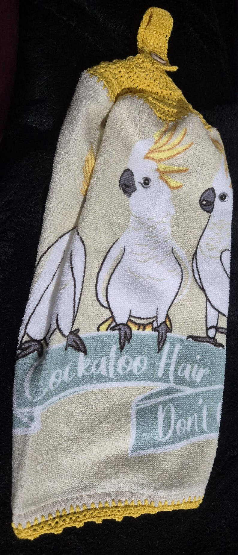 May include: A white and yellow hand towel featuring three cartoon cockatoos with the text "Cockatoo Hair Don't". The towel has a yellow crocheted trim and a loop for hanging.