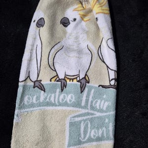 May include: A cream-colored hand towel featuring three cartoon cockatoos. The towel has a light blue banner with the text "Cockatoo Hair Don't Care". The top has a crocheted loop for hanging, and the bottom has a crocheted trim.