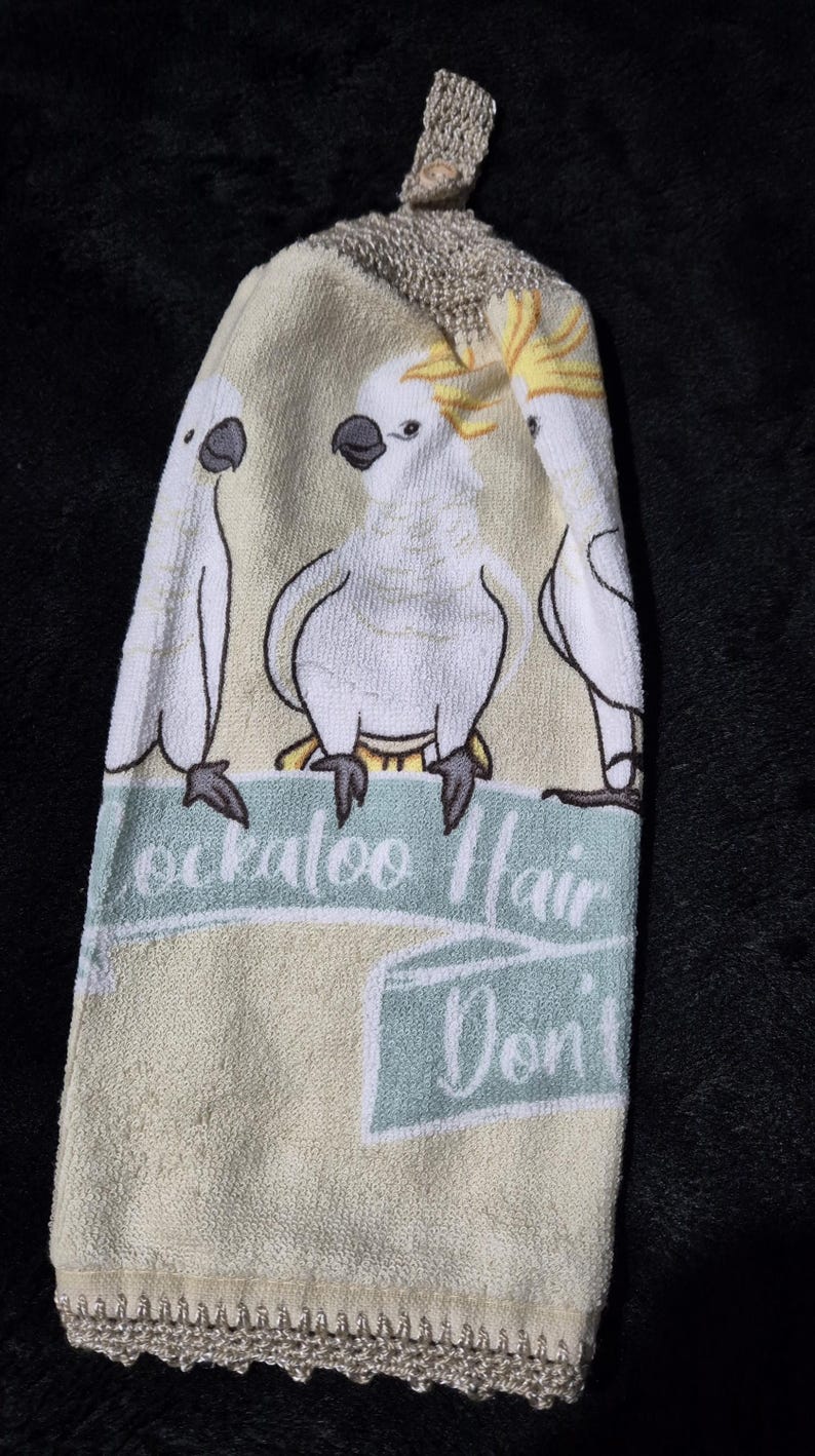 May include: A cream-colored hand towel featuring three cartoon cockatoos. The towel has a light blue banner with the text "Cockatoo Hair Don't Care". The top has a crocheted loop for hanging, and the bottom has a crocheted trim.