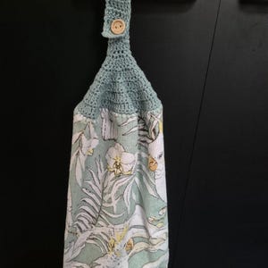 May include: A hanging kitchen towel with a crochet top and a button closure. The towel features a tropical print with white cockatoos, yellow flowers, and green foliage on a light green background.