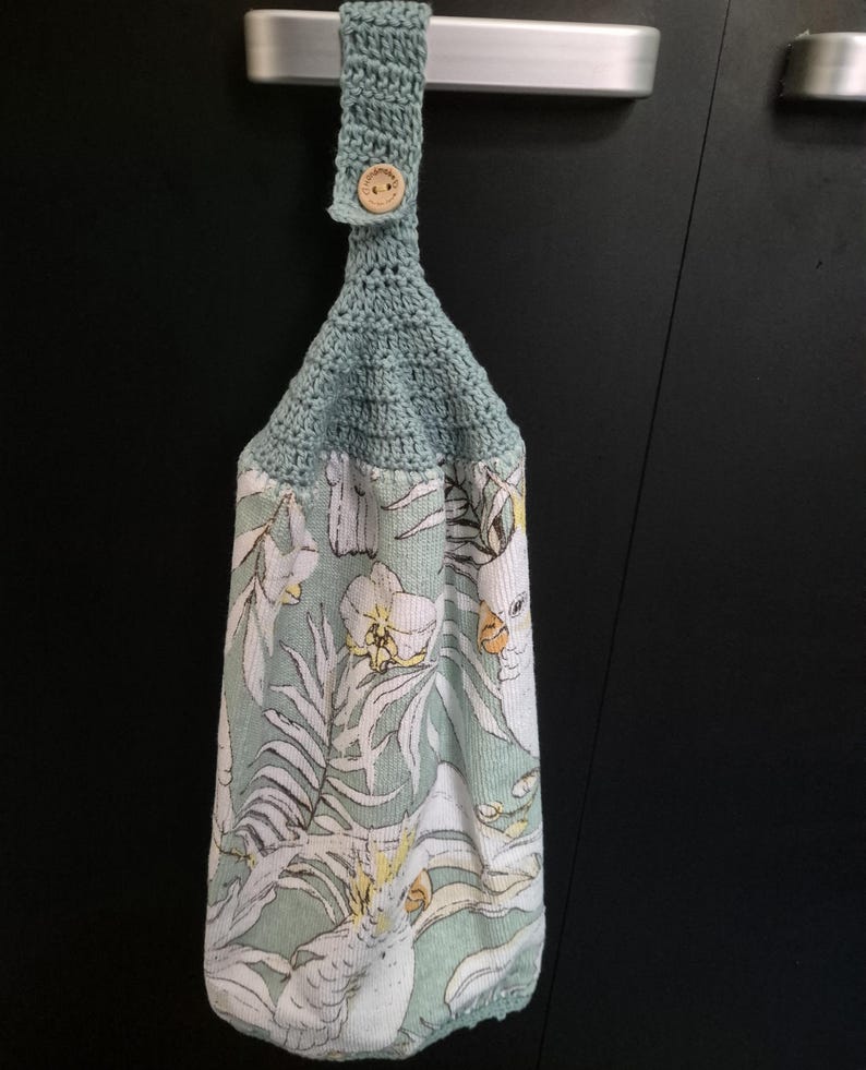 May include: A hanging kitchen towel with a crochet top and a button closure. The towel features a tropical print with white cockatoos, yellow flowers, and green foliage on a light green background.