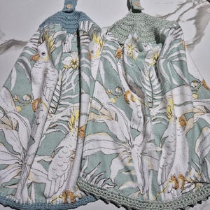 May include: Two hanging kitchen towels with a parrot and floral print. The towels have a light green background with white parrots and flowers. The tops are crocheted in light blue and light green, with a button closure.