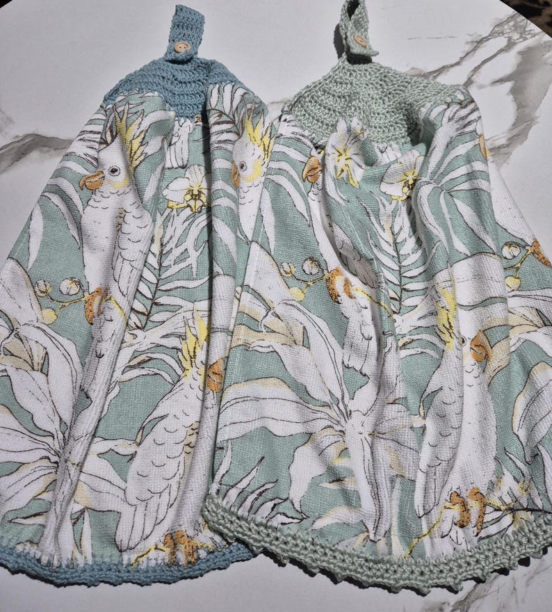 May include: Two hanging kitchen towels with a parrot and floral print. The towels have a light green background with white parrots and flowers. The tops are crocheted in light blue and light green, with a button closure.