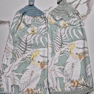 May include: Two hanging kitchen towels with a parrot and floral print. The towels have a light blue and green color scheme with a crocheted top and button closure. The parrot is white with a yellow crest and orange beak.