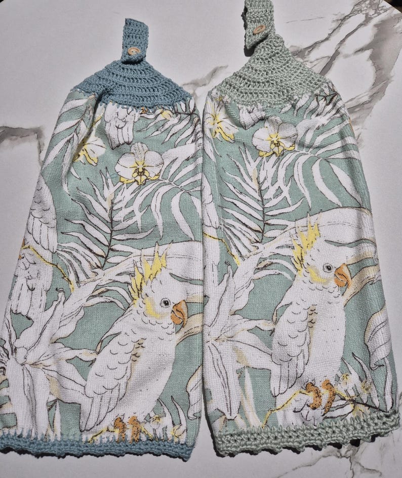 May include: Two hanging kitchen towels with a parrot and floral print. The towels have a light blue and green color scheme with a crocheted top and button closure. The parrot is white with a yellow crest and orange beak.