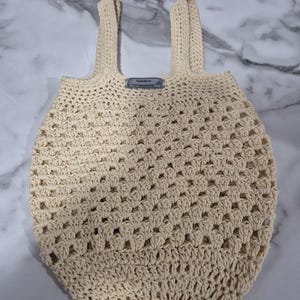 May include: Cream-coloured, handmade crochet tote bag with two sturdy handles. The bag features an open, net-like weave design. A small tag is attached near the top. The bag is sitting on a marble surface.