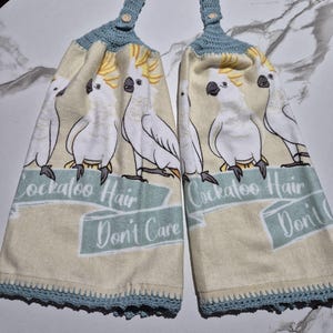 May include: Two beige kitchen towels with a repeating pattern of white cockatoos with yellow crests. The towels have a blue crocheted top and bottom trim. Text on the towels reads "Cockatoo Hair Don't Care".