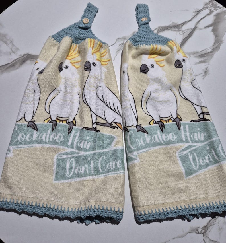 May include: Two beige kitchen towels with a repeating pattern of white cockatoos with yellow crests. The towels have a blue crocheted top and bottom trim. Text on the towels reads "Cockatoo Hair Don't Care".