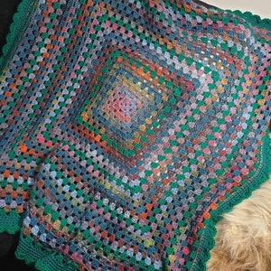 May include: A multi-coloured crochet blanket with a green border and a scalloped edge. The blanket is made with a variety of different coloured yarns, including blue, green, red, orange, and yellow.