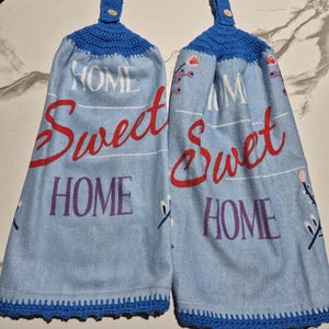 May include: Two blue kitchen towels with the words "HOME Sweet HOME" in red and purple. The towels have a blue crocheted top with a button and a blue crocheted border.