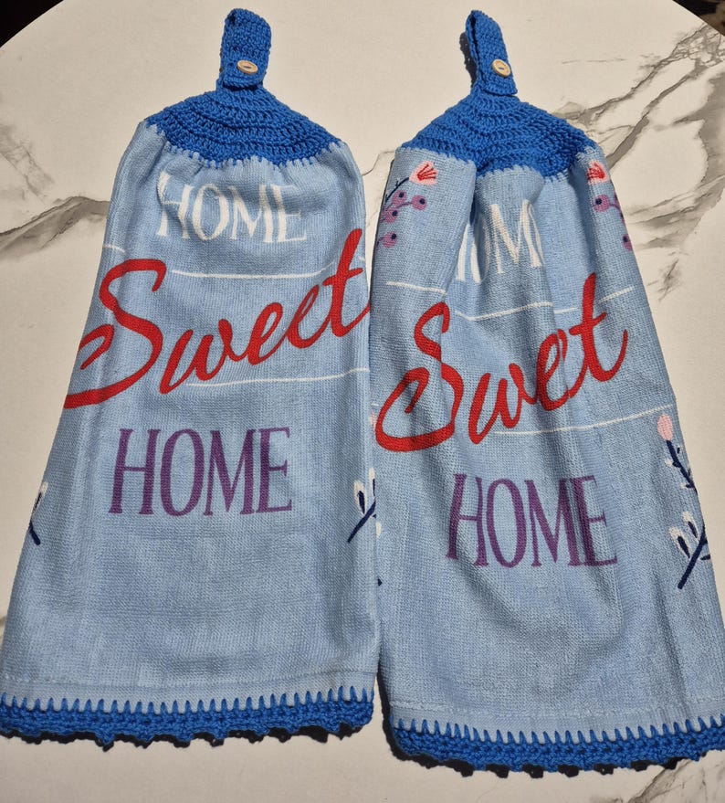 May include: Two blue kitchen towels with the words "HOME Sweet HOME" in red and purple. The towels have a blue crocheted top with a button and a blue crocheted border.